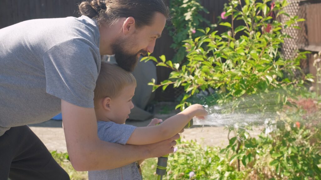 Father and Son. Funny little boy watering lawn plants in garden housing backyard with Dad. Summer house work. Hardworking preschool Kid outdoors. Children help with housework. activity for kids.