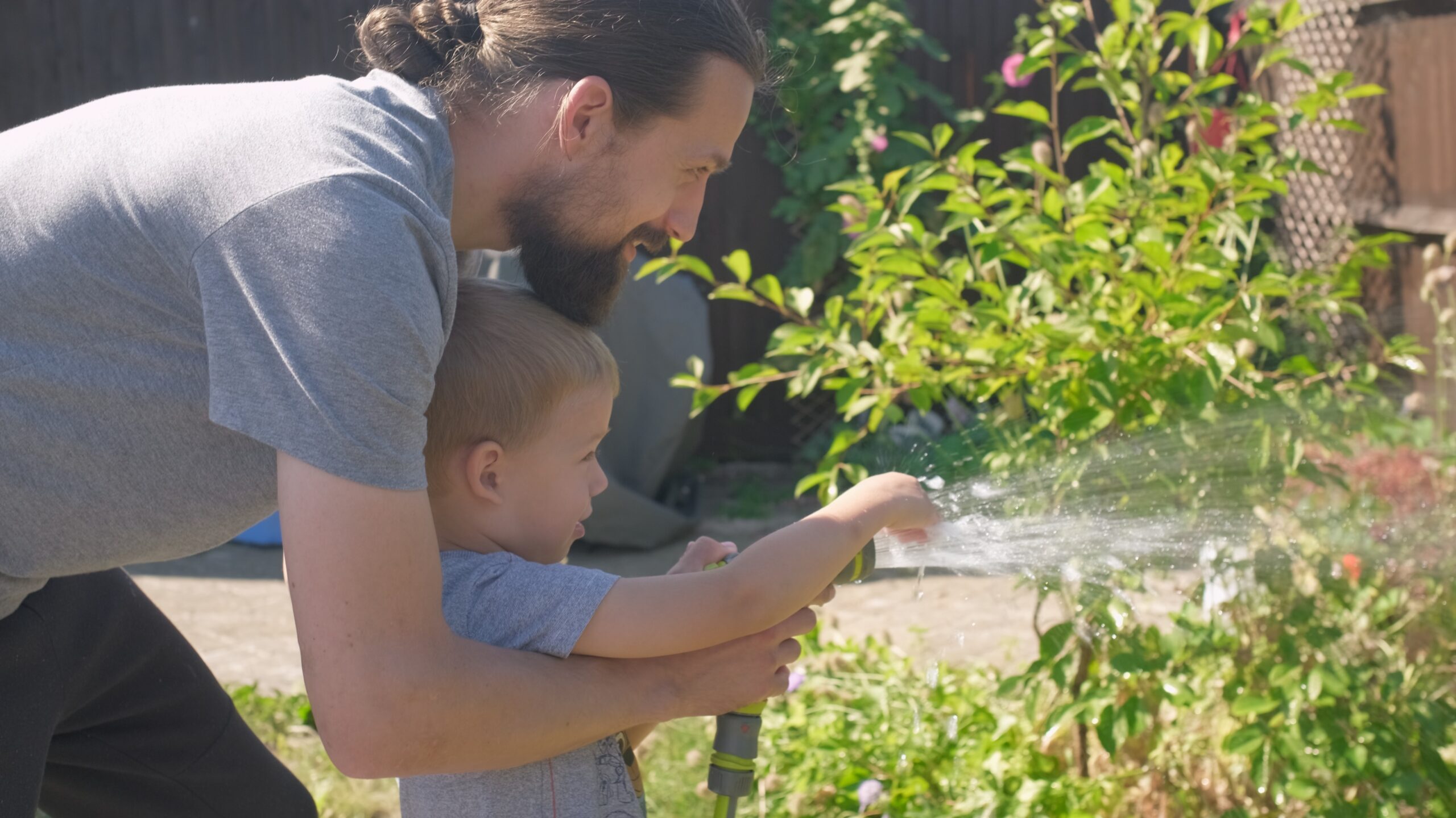 Father and Son. Funny little boy watering lawn plants in garden housing backyard with Dad. Summer house work. Hardworking preschool Kid outdoors. Children help with housework. activity for kids.